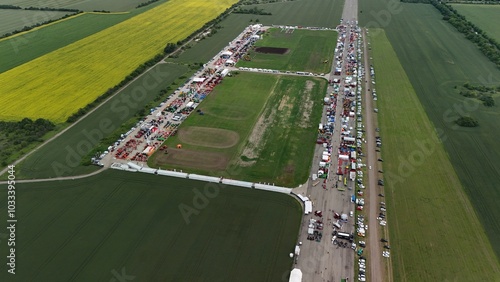This aerial image shows a large agricultural event or trade fair, amidst expansive fields. Multiple rows of tents, machinery, and equipment are neatly arranged on a long track, attracting numerous