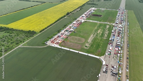This aerial image shows a large agricultural event or trade fair, amidst expansive fields. Multiple rows of tents, machinery, and equipment are neatly arranged on a long track, attracting numerous