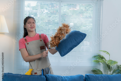 Asian housekeeper in an apron dusts a pillow with a feather duster, ensuring a spotless home. Her focus on cleanliness creates a welcoming, allergen-free living space photo