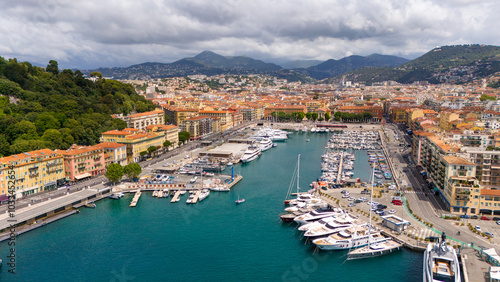 Aerial view of Nice's vibrant port, showcasing luxury yachts docked in turquoise waters. Colorful buildings line the waterfront, with lush green hills and a picturesque cityscape in the background