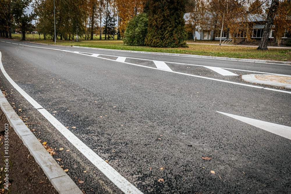 Obraz premium A quiet, empty asphalt road with a white directional arrow and painted lane markings. The scene shows fall foliage and trees in the background, with a nearby building partially visible.