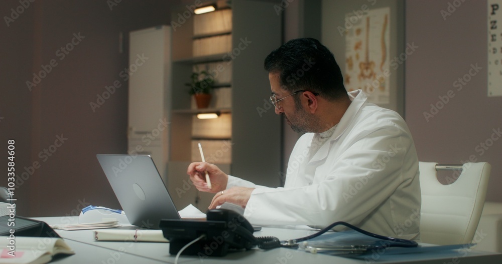 The doctor is typing on a laptop and answering the phone sitting at the table in the medical office, working in the clinic. The doctor is typing on a laptop and answering the phone sitting at the table in the medical office, working in the clinic.
