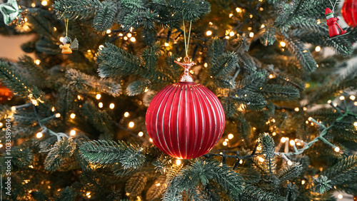 Christmas red ornament close-up hanging on beautifully lit Christmas tree flickering light. Festive bauble is surrounded by glowing string lights pine branches, creating warm cozy holiday atmosphere.