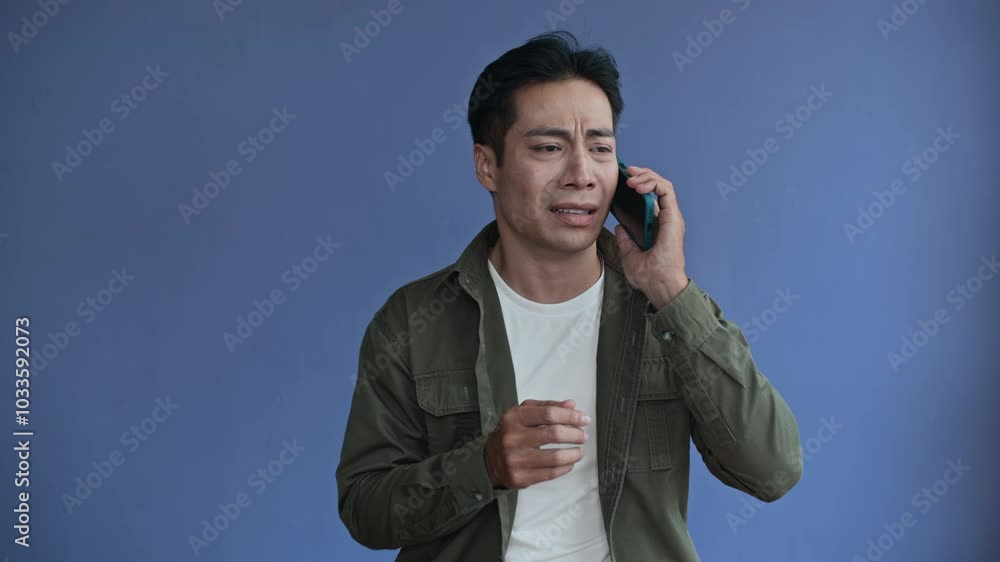Medium copy space studio shot of Asian man being scared and getting bad news from his boss while talking on mobile phone isolated on blue background Medium copy space studio shot of Asian man being scared and getting bad news from his boss while talking on mobile phone isolated on blue background