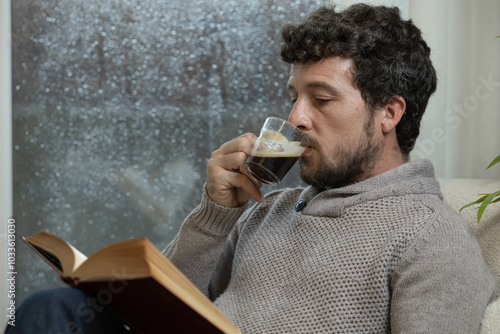 Young adult man enjoys reading a book and having a cup of coffee sitting in an armchair in his living room at home next to the window on a rainy day, warm at home in autumn or winter, reading