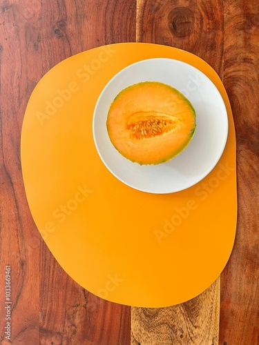 Half a cantaloupe melon on a white plate with an orange placemat, placed on a wooden table, offering a fresh, minimalist food presentation with vibrant colors.