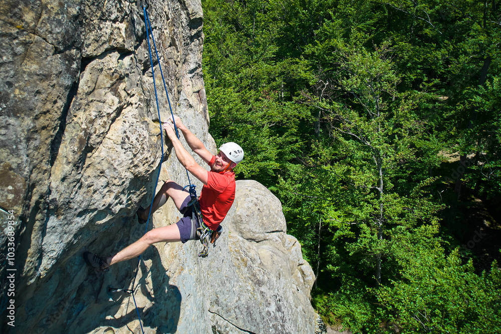 Obraz premium Aerial view of male rock climber ascending rugged limestone cliff with harness and rope for safety. Sportsman climbing on vertical large boulder at Dobvush Rocks in Carpathian mountains, Ukraine.