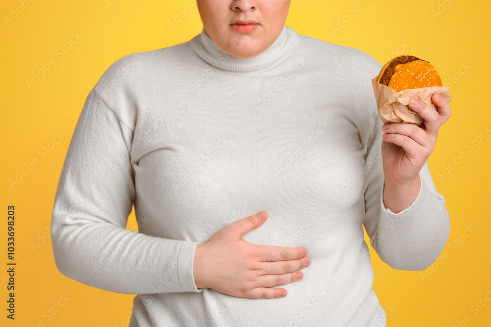 Negative Effects of Fast Food Concept. Plump Girl Holding Burger And Touching Abdomen, Having Stomach Ache, Yellow Studio Background, Copy Space Negative Effects of Fast Food Concept. Plump Girl Holding Burger And Touching Abdomen, Having Stomach Ache, Yellow Studio Background, Copy Space