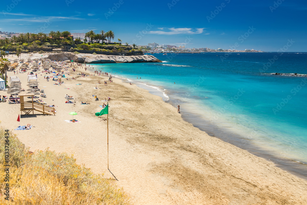 Fototapeta premium A sunny scene at Playa del Duque, Tenerife, with swimmers and sunbathers fill the beach, with a green flag signaling safe swimming