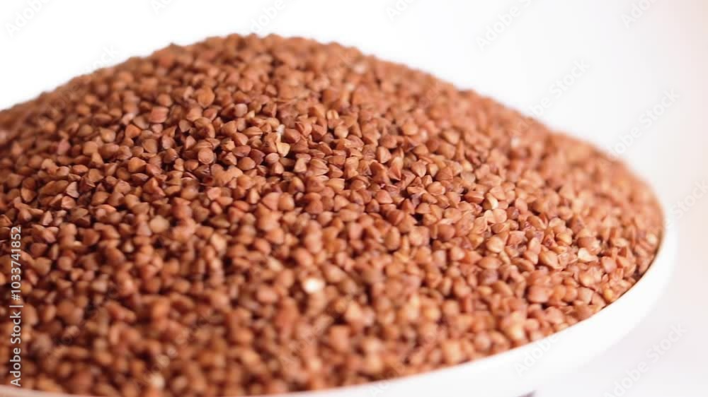 Dry Uncooked Brown Buckwheat Groats Heap on White Plate Rotating against White Background. Raw Large Buckwheat Grains. Russian, Ukrainian and Belarusian Culture Dry Uncooked Brown Buckwheat Groats Heap on White Plate Rotating against White Background. Raw Large Buckwheat Grains. Russian, Ukrainian and Belarusian Culture