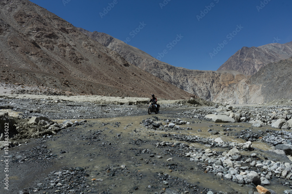 Motorcyclist crossing a rocky riverbed surrounded by rugged mountains Motorcyclist crossing a rocky riverbed surrounded by rugged mountains