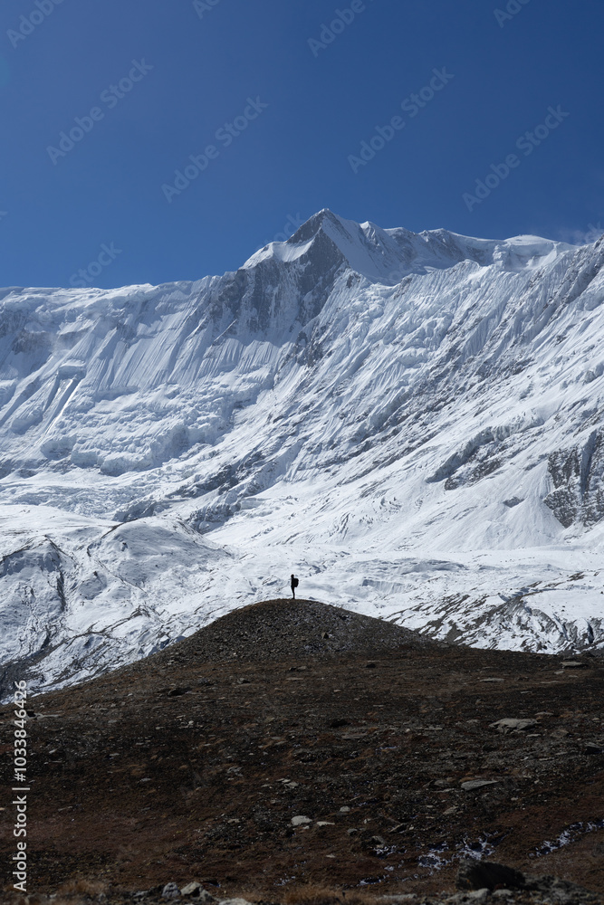 Fototapeta premium A lone hiker stands on a hill, gazing at a majestic snow-covered mountain range under a clear blue sky.