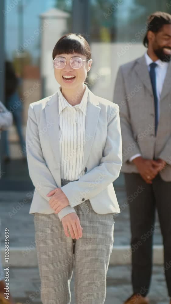 Vertical shot of successful Asian business female leader standing with her multiracial business team in background outside of the building office looking at camera and smiling Vertical shot of successful Asian business female leader standing with her multiracial business team in background outside of the building office looking at camera and smiling