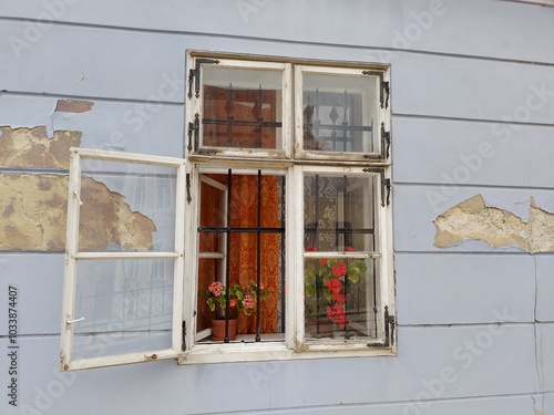 Open window sash at old blue painted house in citycenter of Győr, Hungary. Decorative flowers in front of grid. Patterned orange thick curtain, white light lace curtain. Waiting for summer breeze. photo