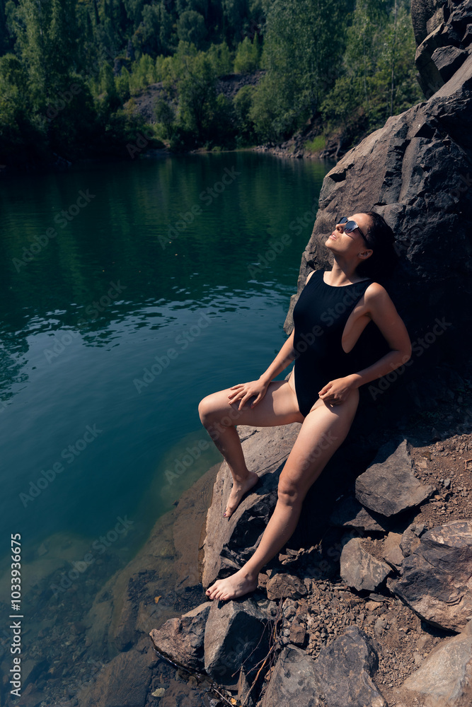 Relaxing by Serene Lake in Nature, Woman in Black Swimsuit Enjoying Sun on Rocky Shore Relaxing by Serene Lake in Nature, Woman in Black Swimsuit Enjoying Sun on Rocky Shore