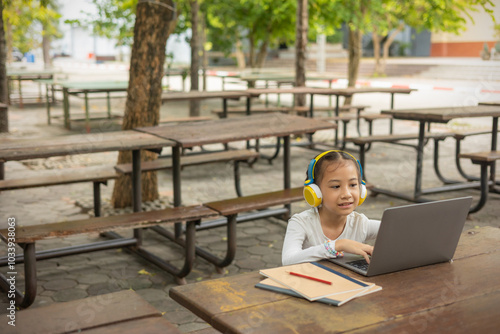 Focused Asian child engages in online learning outdoors, using a laptop and headphones. It shows the role of technology and digital devices in remote education in modern learning environments.