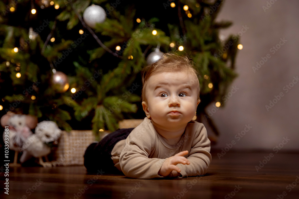Curious baby lies on floor in front of decorated christmas tree adorned with lights and ornaments. cozy ambiance highlights child's inquisitive expression during festive season. Curious baby lies on floor in front of decorated christmas tree adorned with lights and ornaments. cozy ambiance highlights child's inquisitive expression during festive season.