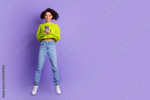 Photo of curly american youngster jumping girl in jeans and sweater using mobile phone isolated on purple color background