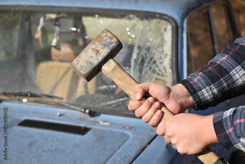 A man is clutching a hammer in front of a bright blue car