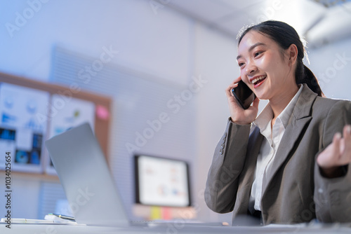 Smiling young businesswoman multitasks with phone and laptop in office. Confidence and happiness shine as she navigates tech and communication