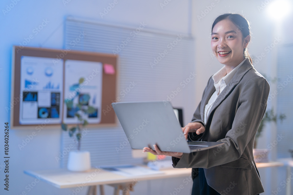 Confident asian businesswoman works late in the office, smiling happily at her laptop screen, using modern technology to manage work efficiently and strategize for success Confident asian businesswoman works late in the office, smiling happily at her laptop screen, using modern technology to manage work efficiently and strategize for success