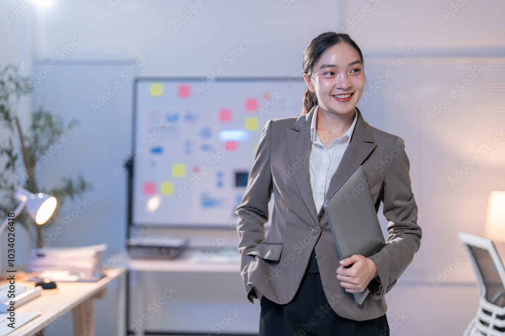 Young manager woman is holding a laptop and smiling while standing in an office at night. She is wearing a suit and is confident in her work Young manager woman is holding a laptop and smiling while standing in an office at night. She is wearing a suit and is confident in her work