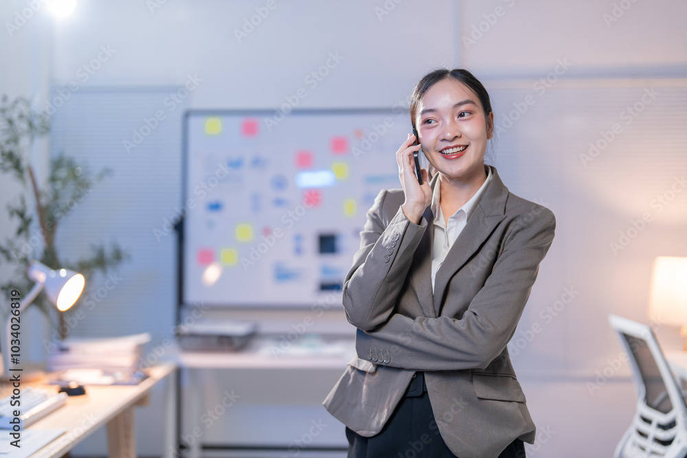 Businesswoman is standing in her office at night, smiling while talking on her phone and going over notes on a whiteboard Businesswoman is standing in her office at night, smiling while talking on her phone and going over notes on a whiteboard