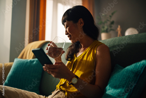 Asian woman in yellow top reclines on teal sofa, drinking water, phone near. photo