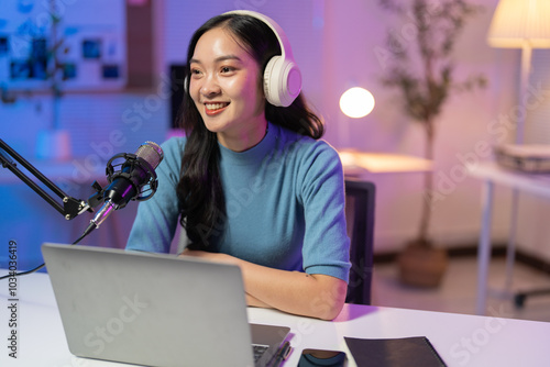 Young asian woman is recording a podcast from home, talking into a microphone and using a laptop. She is smiling and looking away from the camera photo