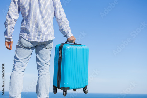 Happy single young adult man on his back with his blue travel suitcase with the background of blue sky and ocean or sea, resting vacation, traveling the world, traveling alone, having fun, hotel booki