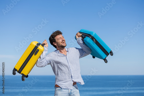 Happy and euphoric single young adult man with his yellow and blue travel suitcases with the background of blue sky and ocean or sea, resting vacation, traveling the world, traveling alone, having fun