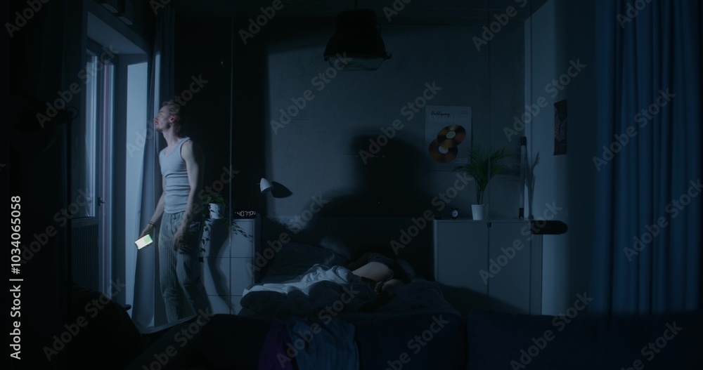 A man uses a mobile phone while walking around the bedroom at night. His wife is sleeping in bed. Dark room, home interior A man uses a mobile phone while walking around the bedroom at night. His wife is sleeping in bed. Dark room, home interior