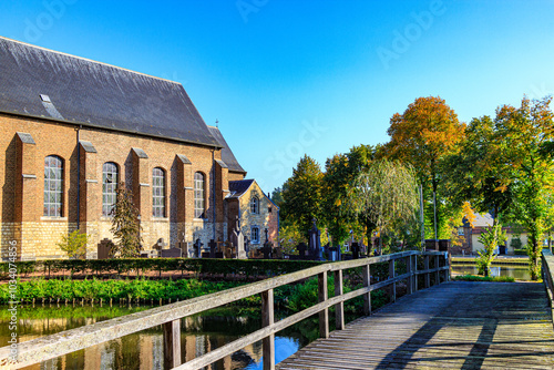 Wooden bridge over castle moat, brick walls of church, cemetery, public park with pond and autumn trees in background, sunny day in Wijnandsrade, South Limburg, Netherlands photo