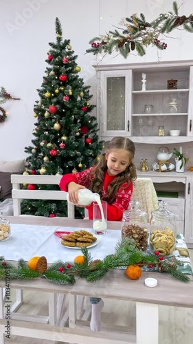 Happy little girl in red sweater pouring milk from a bottle into a glass in the kitchen on Christmas, vertical footage. Cute child drinking milk near Christmas tree. Christmas holidays.