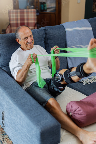 An elderly man is using a resistance band to perform vital rehabilitation exercises within the comfort of home. Tough exercises, pain during recovery. Indoors. Vertical