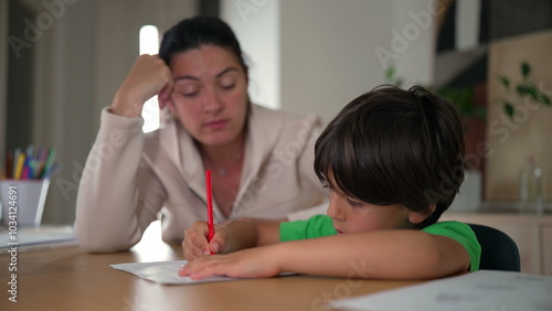 Mother and young boy working on homework together at a table. mother looks tired and bored, while the boy is focused and concentrating on his task in a home setting