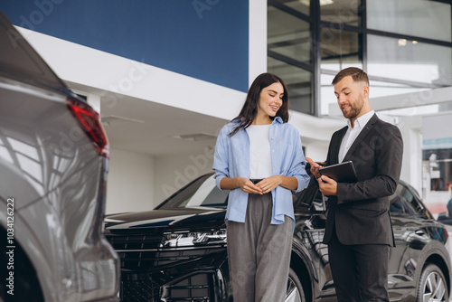 Young woman customer female buyer client wears shirt consult with salesman hold tablet choosing auto want to buy new automobile in car showroom vehicle salon dealership store motor show.