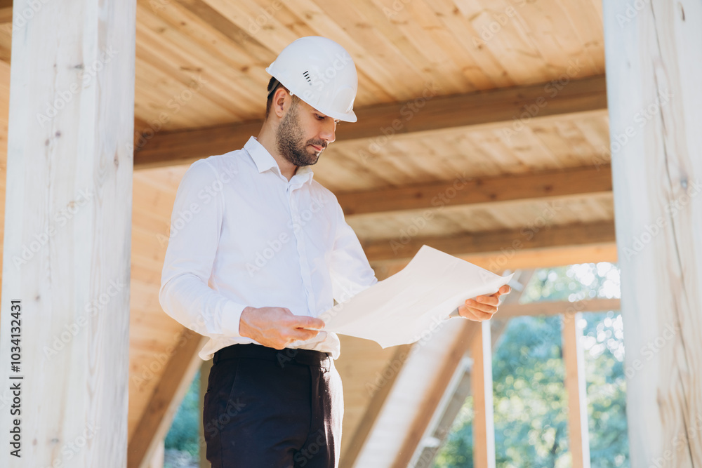 Construction engineer in white helmet. Developer with construction documentation on the background of wooden modular building under construction. Portrait of smiling architect near eco friendly house. Construction engineer in white helmet. Developer with construction documentation on the background of wooden modular building under construction. Portrait of smiling architect near eco friendly house.