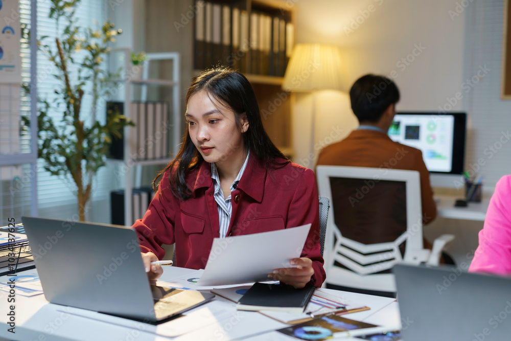 Young asian businesswoman analyzing paperwork and working on laptop computer with colleagues in background Young asian businesswoman analyzing paperwork and working on laptop computer with colleagues in background