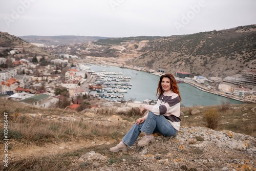 A woman is sitting on a rock overlooking a body of water. The scene is peaceful and serene, with the woman looking out over the water and taking in the view.