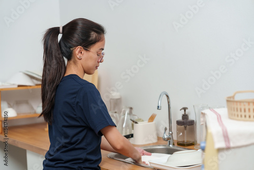 Asian woman washing dishes at sink in kitchen with focused expression. Long black hair tied back, wearing glasses. Neatly organized kitchen counter with dish rack, glasses, and plates. clean setting.