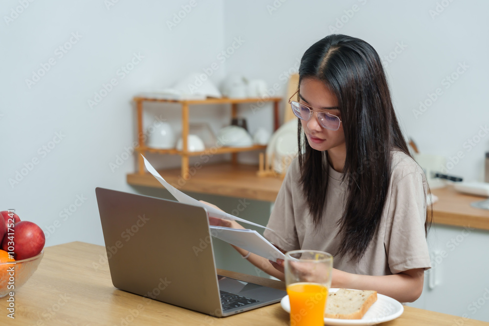 Asian woman working at kitchen table reviewing printed documents. Focused expression. Laptop, fruit bowl, and breakfast items placed on wooden counter. Multitasking during morning routine Asian woman working at kitchen table reviewing printed documents. Focused expression. Laptop, fruit bowl, and breakfast items placed on wooden counter. Multitasking during morning routine