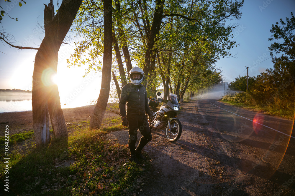 A motorcyclist is enjoying the last warm days of autumn. Trees and sun rays of sunset. the road near the lake. A biker in dark special clothes and a white dual sport helmet. Silhouette of a motorcycle