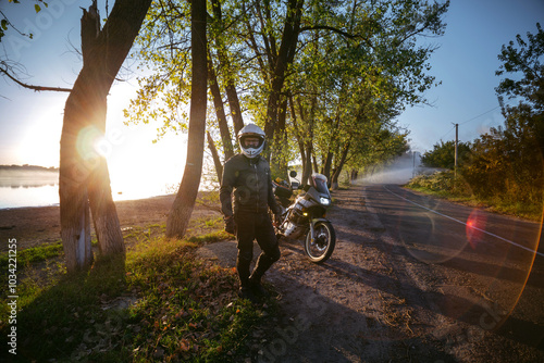Obraz na płótnie A motorcyclist is enjoying the last warm days of autumn. Trees and sun rays of sunset. the road near the lake. A biker in dark special clothes and a white dual sport helmet. Silhouette of a motorcycle w salonie