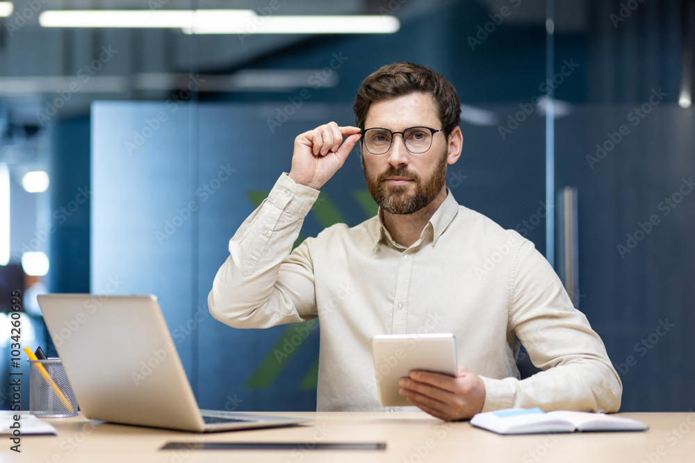 Portrait of a young serious businessman sitting in the office at the desk, holding a tablet in his hand and wearing glasses, confidently looking at the camera Portrait of a young serious businessman sitting in the office at the desk, holding a tablet in his hand and wearing glasses, confidently looking at the camera