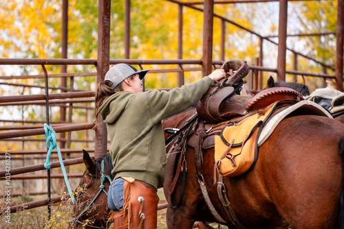 cowgirl wearing hat and sweatshirt saddling a horse with western saddle and tightening cinch at corrals in mountains photo