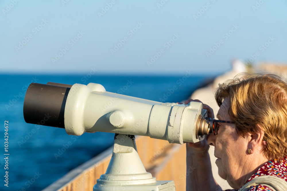Obraz premium An older woman looking at the sea through binoculars at a viewpoint.