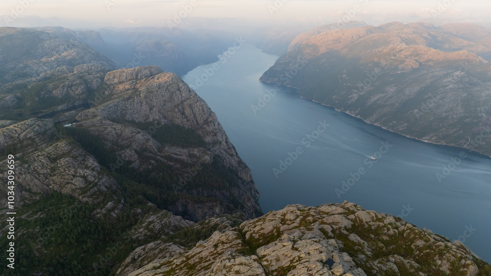 Aerial shot of a couple standing at the edge of Preikestolen. First morning sunbeams reach the cliff formation. Massive rocks surrounding the couple. Dangerous place for a hike. Beauty of the nature Aerial shot of a couple standing at the edge of Preikestolen. First morning sunbeams reach the cliff formation. Massive rocks surrounding the couple. Dangerous place for a hike. Beauty of the nature