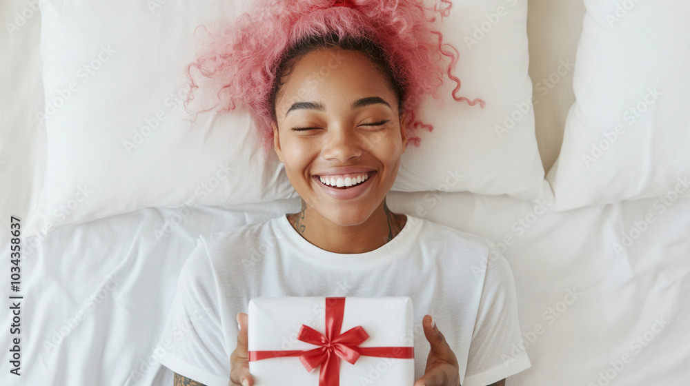 A young woman with pink hair smiles joyfully while holding a gift box on a bed with white pillows in a bright room, happy valentine's day