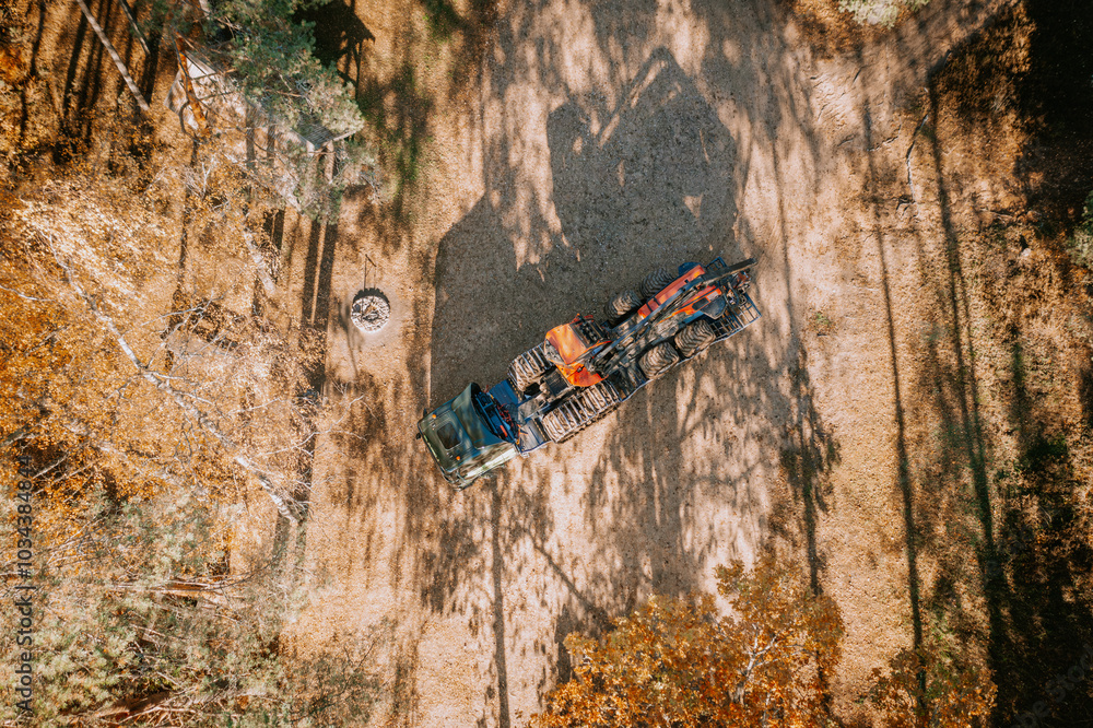 Obraz premium Aerial shot showing a truck with logging machinery on a trailer in a forest clearing, surrounded by trees and shadows on the ground.